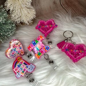 May include: A collection of pink and colorful badge reels and a keychain. The badge reels feature heart and rainbow shapes with the phrase "Mañana Será Bonito" in various colors. The keychain has a pink heart with the same phrase and a pink tassel.