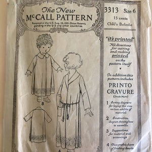 Op de afbeelding: Vintage McCall-patroon voor een kinderbadjas. De patroonhoes bevat illustraties van het badjasontwerp, samen met tekst met instructies en kenmerken. De envelop is crèmekleurig met zwarte tekst en illustraties.