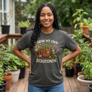 May include: Dark gray t-shirt with the text "I GROW MY OWN SEROTONIN". The shirt features a graphic of a basket filled with vegetables, flowers, and gardening tools. The person is smiling and standing in a garden.