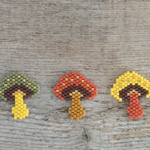 May include: Three beaded mushroom charms in different colors. The mushrooms are made with brown, green, yellow, orange, and gold beads. The mushrooms are arranged in a row on a wooden surface.