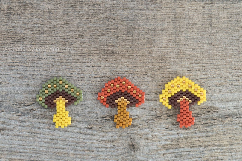 May include: Three beaded mushroom charms in different colors. The mushrooms are made with brown, green, yellow, orange, and gold beads. The mushrooms are arranged in a row on a wooden surface.