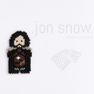 Op de afbeelding: Een zwart-witte pixel art kralen-sprite van Jon Snow uit Game of Thrones. Het personage draagt een zwarte mantel en een bruine riem. De tekst "Jon Snow" staat in wit boven het personage. Een grijs-witte pixel art kralen-sprite van een direwolf bevindt zich aan de rechterkant van de afbeelding.