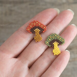 May include: Two beaded mushroom charms, one with a red and orange cap and the other with a green cap. Both have brown stems and yellow accents.