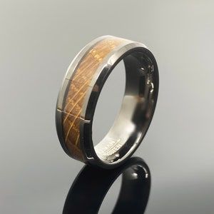 May include: A black tungsten ring with a brown wood inlay. The ring has a beveled edge and a polished finish. The inscription "TUNGSTEN" is visible on the inside of the band.