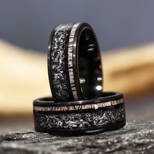 Puede incluir: Dos anillos negros con un diseño único. Los anillos presentan una banda de material blanco y marrón, con una incrustación texturizada gris oscuro. Los anillos están apilados, mostrando sus intrincados detalles.