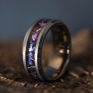 Puede incluir: Un anillo oscuro con una incrustación de galaxia púrpura, azul y blanca. El anillo tiene una banda oscura con un borde plateado. El anillo se encuentra sobre un fondo borroso, resaltando el diseño y los colores.