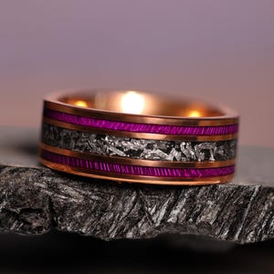 May include: A rose gold ring with a striking design. The ring features a band of vibrant magenta and a center section of textured, silver-toned material. The ring is set on a dark, textured surface.