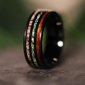 May include: A black ring with a unique design. The ring features a band of brown and white material, a green band, and a band of reddish-brown wood. The ring is set against a blurred background.