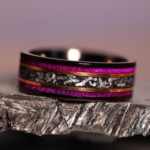 May include: A black ring with a vibrant magenta inlay and copper-colored bands. The center band features a textured, silver-toned material. The ring is displayed on a textured, gray surface.