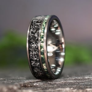May include: A close-up of a black and silver ring with a unique design. The ring features a dark, textured band with a central inlay of green and white stone. The ring's polished silver edges provide a striking contrast to the darker elements.