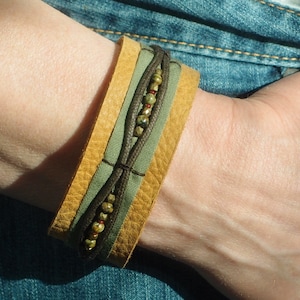Op de afbeelding: Een bruin leren armband met een groene stoffen band en een rij gekleurde kralen.