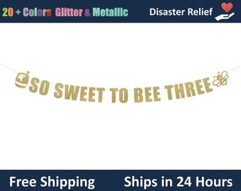 May include: A gold glitter banner with the phrase "SO SWEET TO BEE THREE" in capital letters. The banner includes a honey pot graphic on the left and a bee graphic on the right. The banner is strung on a thin white string.