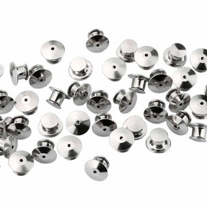 May include: A collection of shiny, silver-colored pin backs, also known as butterfly clutches, scattered against a white background. These small, round fasteners have a post and a flat, disc-shaped back, designed to secure pins.