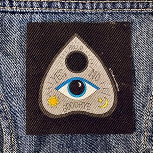 May include: A black and white ouija board with a blue eye, a sun, a moon, and stars. The board has the words "yes", "no", "hello", "goodbye", and "I love you" written on it. The board is on a blue denim background.