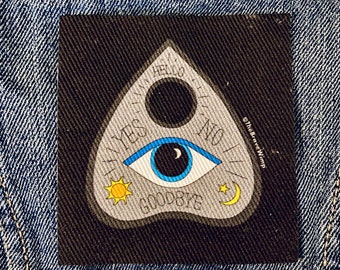 Puede incluir: Una tabla de ouija en blanco y negro con un ojo azul, un sol, una luna y estrellas. La tabla tiene las palabras "sí", "no", "hola", "adiós" y "te amo" escritas en ella. La tabla está sobre un fondo de mezclilla azul.