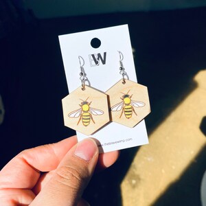 May include: A pair of wooden earrings shaped like hexagons with a bee design. The bees are yellow and black with a white background. The earrings are on a white card with the text "www.thebravewimp.com".