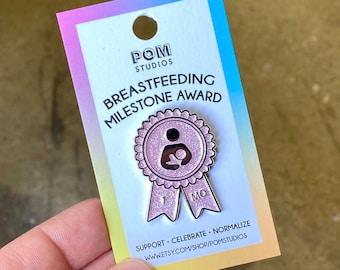 May include: Pink glitter enamel pin with a ribbon and the text "1 MO" on it. The pin features a stylized image of a person breastfeeding a baby. The text "BREASTFEEDING MILESTONE AWARD" is printed on the card behind the pin. The text "SUPPORT. CELEBRATE. NORMALIZE" is printed on the bottom of the card. The text "WWW.ETSY.COM/SHOP/POMSTUDIOS" is printed below the previous text.