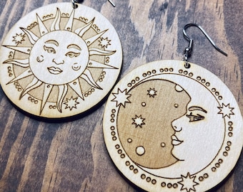 May include: A pair of wooden earrings featuring a sun and moon design. The sun and moon are both stylized with faces and surrounded by stars. The earrings are attached to silver hooks.
