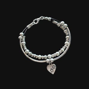 May include: A silver bracelet with a heart charm featuring paw prints. The bracelet is made of two strands of silver beads and a black cord.