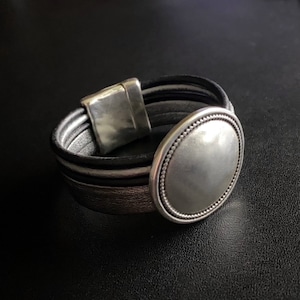 May include: A silver and brown leather bracelet with a large, round, silver-toned charm. The charm has a beaded edge and a smooth, flat surface.