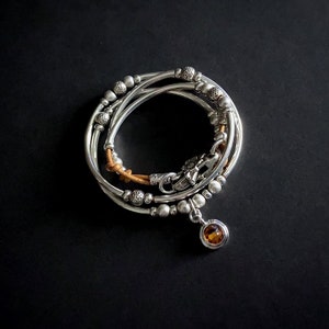 May include: A silver bracelet with a brown leather strap and a small amber gemstone charm. The bracelet is made of multiple strands of silver beads.