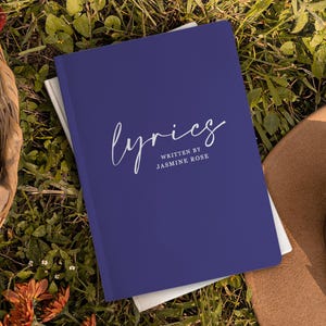 May include: A dark blue notebook with the word "lyrics" written in white cursive script. The text "written by Jasmine Rose" is printed in white below the word "lyrics".