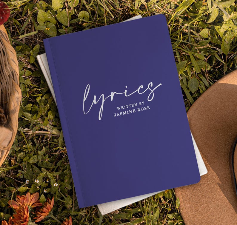 May include: A dark blue notebook with the word "lyrics" written in white cursive script. The text "written by Jasmine Rose" is printed in white below the word "lyrics".