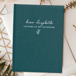 Peut inclure: Un journal de couleur teal avec le texte "dear elizabeth LETTERS TO MY DAUGHTER" en &eacute;criture blanche. Une petite illustration florale est en dessous du texte. Le journal est sur une enveloppe blanche et une surface &agrave; motifs or et blanc.