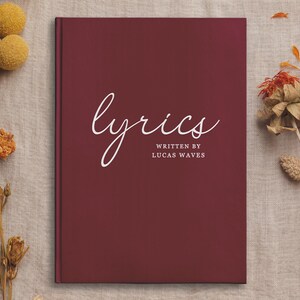 May include: A burgundy hardcover journal with the word "lyrics" written in white cursive script. The text "written by Lucas Waves" is printed in white below the word "lyrics".