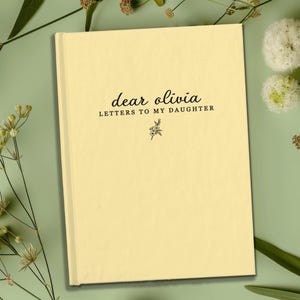 Peut inclure: Un journal cr&egrave;me avec le texte "dear olivia LETTERS TO MY DAUGHTER" en &eacute;criture noire. Une petite illustration florale se trouve sous le texte. Le journal est entour&eacute; de feuillage vert et de fleurs blanches.