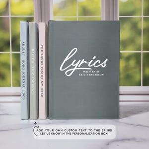 May include: Three gray hardcover journals with white text on the spines. The journal on the right has the text "lyrics" with "written by Eric Henderson" below it. The middle journal has the text "The Songs Inside My Head". The left journal has the text "Daisy's Lyric Journal". The text on the leftmost journal is partially obscured by the other journals. The text "Add your own custom text to the spine! Let us know in the personalization box!" is below the journals.