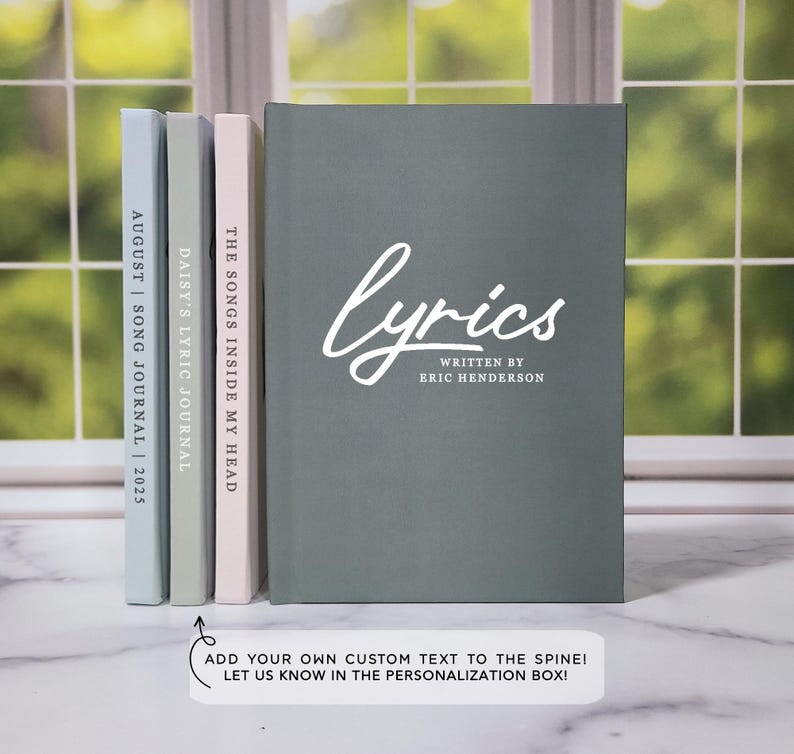 May include: Three gray hardcover journals with white text on the spines. The journal on the right has the text "lyrics" with "written by Eric Henderson" below it. The middle journal has the text "The Songs Inside My Head". The left journal has the text "Daisy's Lyric Journal". The text on the leftmost journal is partially obscured by the other journals. The text "Add your own custom text to the spine! Let us know in the personalization box!" is below the journals.