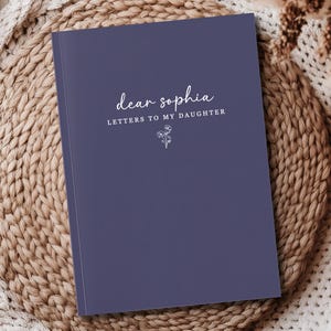 Peut inclure: Un journal bleu marine avec le texte "dear sophia LETTERS TO MY DAUGHTER" en &eacute;criture blanche. Une petite illustration florale est en dessous du texte. Le journal repose sur une surface beige tiss&eacute;e.