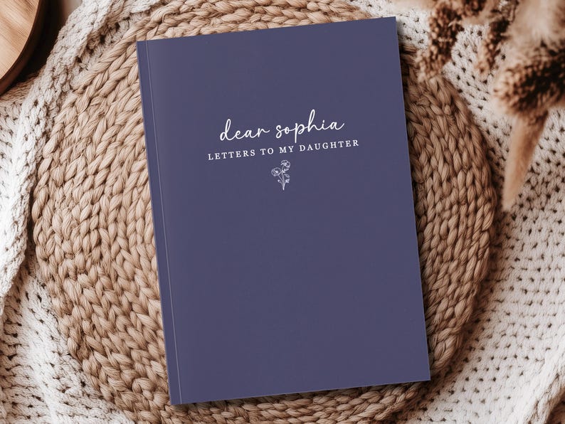 Peut inclure: Un journal bleu marine avec le texte "dear sophia LETTERS TO MY DAUGHTER" en &eacute;criture blanche. Une petite illustration florale est en dessous du texte. Le journal repose sur une surface beige tiss&eacute;e.