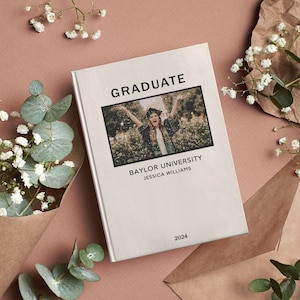 May include: A white book with the word "GRADUATE" in black text. The book has a photo of a person in a graduation cap and gown throwing confetti in the air. The text "BAYLOR UNIVERSITY" and "JESSICA WILLIAMS" is below the photo. The year "2024" is at the bottom of the book.