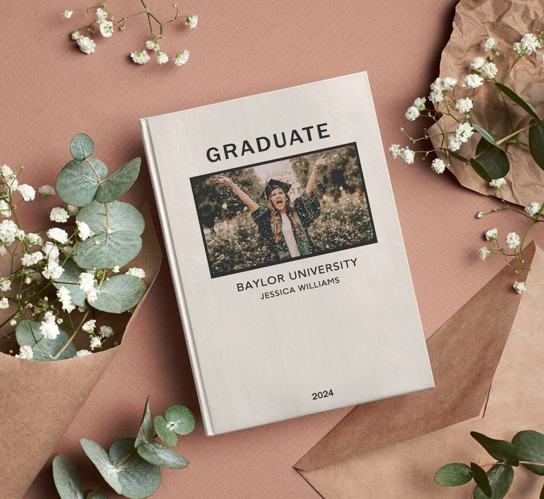 May include: A white book with the word "GRADUATE" in black text. The book has a photo of a person in a graduation cap and gown throwing confetti in the air. The text "BAYLOR UNIVERSITY" and "JESSICA WILLIAMS" is below the photo. The year "2024" is at the bottom of the book.