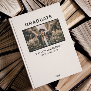 May include: A white book with the word "GRADUATE" in black text on the cover. The book has a photo of a person in a graduation cap and gown throwing confetti in the air. The text "BAYLOR UNIVERSITY" and "JESSICA WILLIAMS" is below the photo. The year "2024" is at the bottom of the cover.