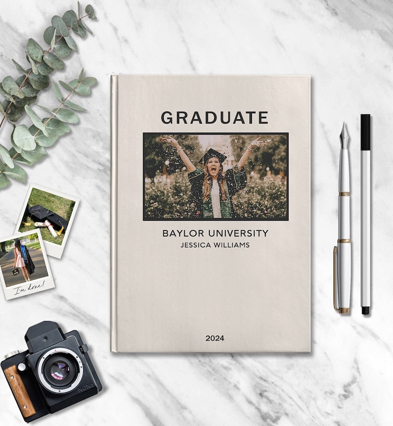 May include: A beige book with the text "GRADUATE" in black lettering. The book features a photo of a person in a graduation cap and gown throwing confetti in the air. The text "BAYLOR UNIVERSITY" and "JESSICA WILLIAMS" is printed below the photo. The year "2024" is printed at the bottom of the book.