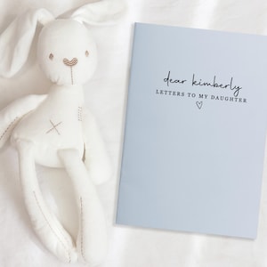Peut inclure: Un lapin en peluche blanc avec de longues oreilles et des d&eacute;tails brod&eacute;s, &agrave; c&ocirc;t&eacute; d'un livre bleu clair. La couverture du livre indique "dear kimberly LETTERS TO MY DAUGHTER" avec un petit symbole de c&oelig;ur.