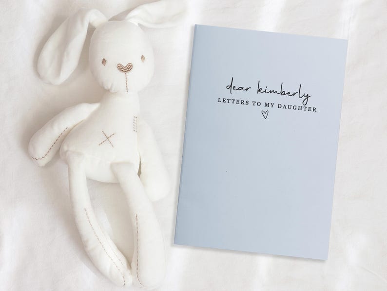 Peut inclure: Un lapin en peluche blanc avec de longues oreilles et des d&eacute;tails brod&eacute;s, &agrave; c&ocirc;t&eacute; d'un livre bleu clair. La couverture du livre indique "dear kimberly LETTERS TO MY DAUGHTER" avec un petit symbole de c&oelig;ur.