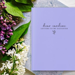 Peut inclure: Un journal lavande avec le texte "dear caroline LETTERS TO MY DAUGHTER" en &eacute;criture noire. Une illustration florale est en dessous du texte. Le journal est &agrave; c&ocirc;t&eacute; d'un bouquet de fleurs violettes et blanches avec des feuilles vertes.