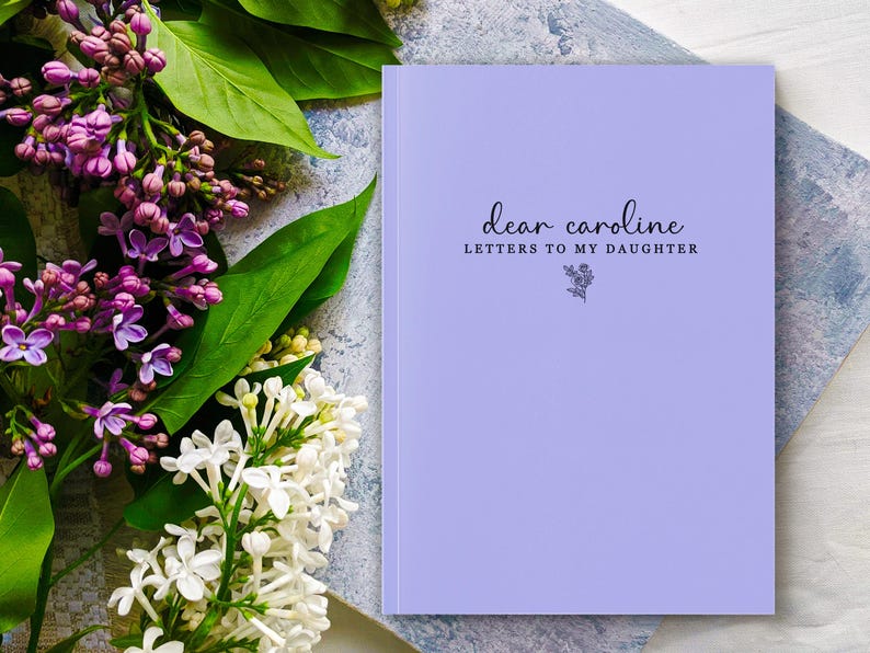 Peut inclure: Un journal lavande avec le texte "dear caroline LETTERS TO MY DAUGHTER" en &eacute;criture noire. Une illustration florale est en dessous du texte. Le journal est &agrave; c&ocirc;t&eacute; d'un bouquet de fleurs violettes et blanches avec des feuilles vertes.