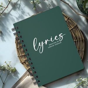 May include: A dark green spiral bound notebook with the word "lyrics" written in white cursive script. The text "written by Leslie Jordan" is printed in white below the word "lyrics".