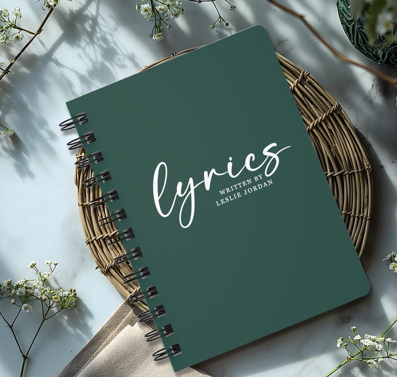 May include: A dark green spiral bound notebook with the word "lyrics" written in white cursive script. The text "written by Leslie Jordan" is printed in white below the word "lyrics".
