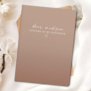 Peut inclure: Un journal &agrave; couverture rigide marron clair avec le texte "dear madison LETTERS TO MY DAUGHTER" &eacute;crit en script. Un petit c&oelig;ur est en dessous du texte. Des fleurs blanches et un tissu blanc en arri&egrave;re-plan.