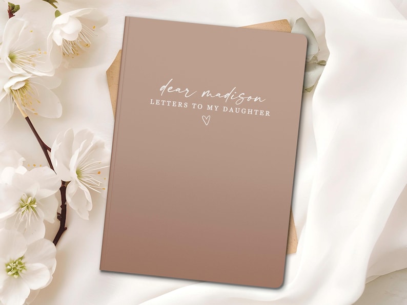Peut inclure: Un journal &agrave; couverture rigide marron clair avec le texte "dear madison LETTERS TO MY DAUGHTER" &eacute;crit en script. Un petit c&oelig;ur est en dessous du texte. Des fleurs blanches et un tissu blanc en arri&egrave;re-plan.