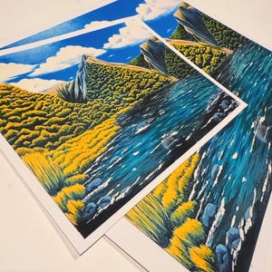 Puede incluir: Tres impresiones art&iacute;sticas que presentan una escena de paisaje vibrante. La obra de arte representa un cielo azul con nubes blancas, una ladera amarilla y verde y un r&iacute;o azul. Las impresiones est&aacute;n sobre papel blanco y se muestran en &aacute;ngulo.