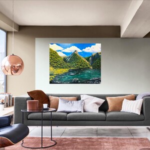 Puede incluir: Una pintura de paisaje representa una cordillera con exuberante vegetaci&oacute;n verde, un lago sereno y un cielo azul brillante con nubes blancas y esponjosas. La obra de arte se exhibe sobre un sof&aacute; gris en una sala de estar moderna.