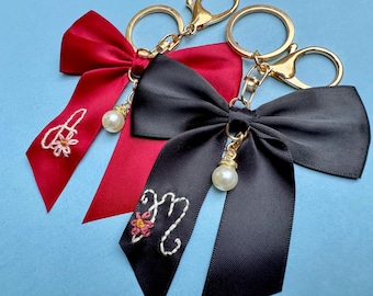 Peut inclure: Trois porte-clés avec des nœuds en ruban de satin rouge et noir. Chaque nœud est orné d'un fermoir doré et d'une fausse perle pendante. Le nœud rouge porte des détails floraux brodés et la lettre "A", tandis que le nœud noir porte la lettre "M".