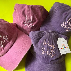 Puede incluir: Cuatro gorras de béisbol de color rosa y morado con texto bordado. Las gorras dicen "big sis", "lil sis" y "big sis".